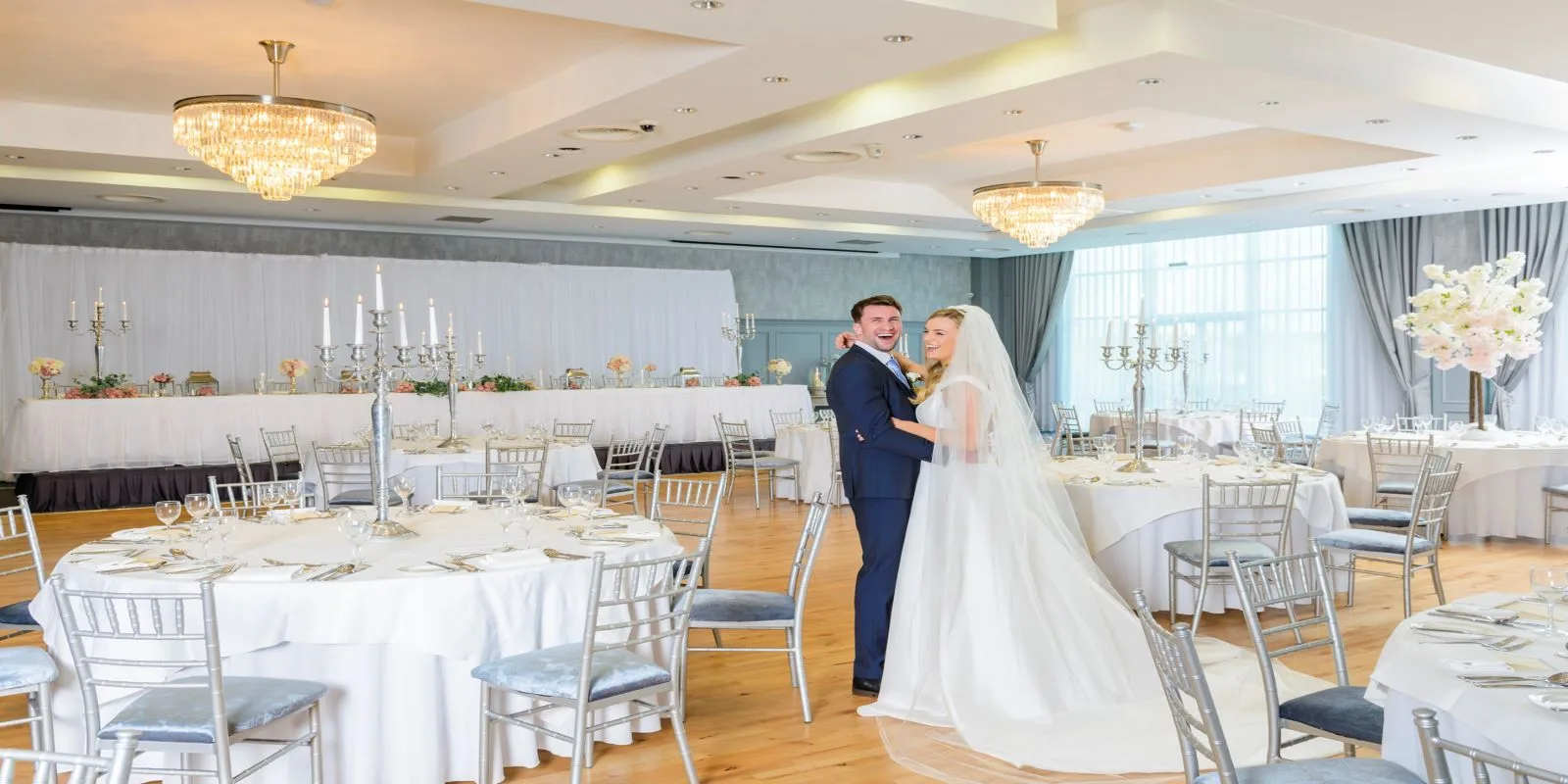 Representative hotel space: A wedding couple poses in a bright banquet hall with round tables and chandeliers.