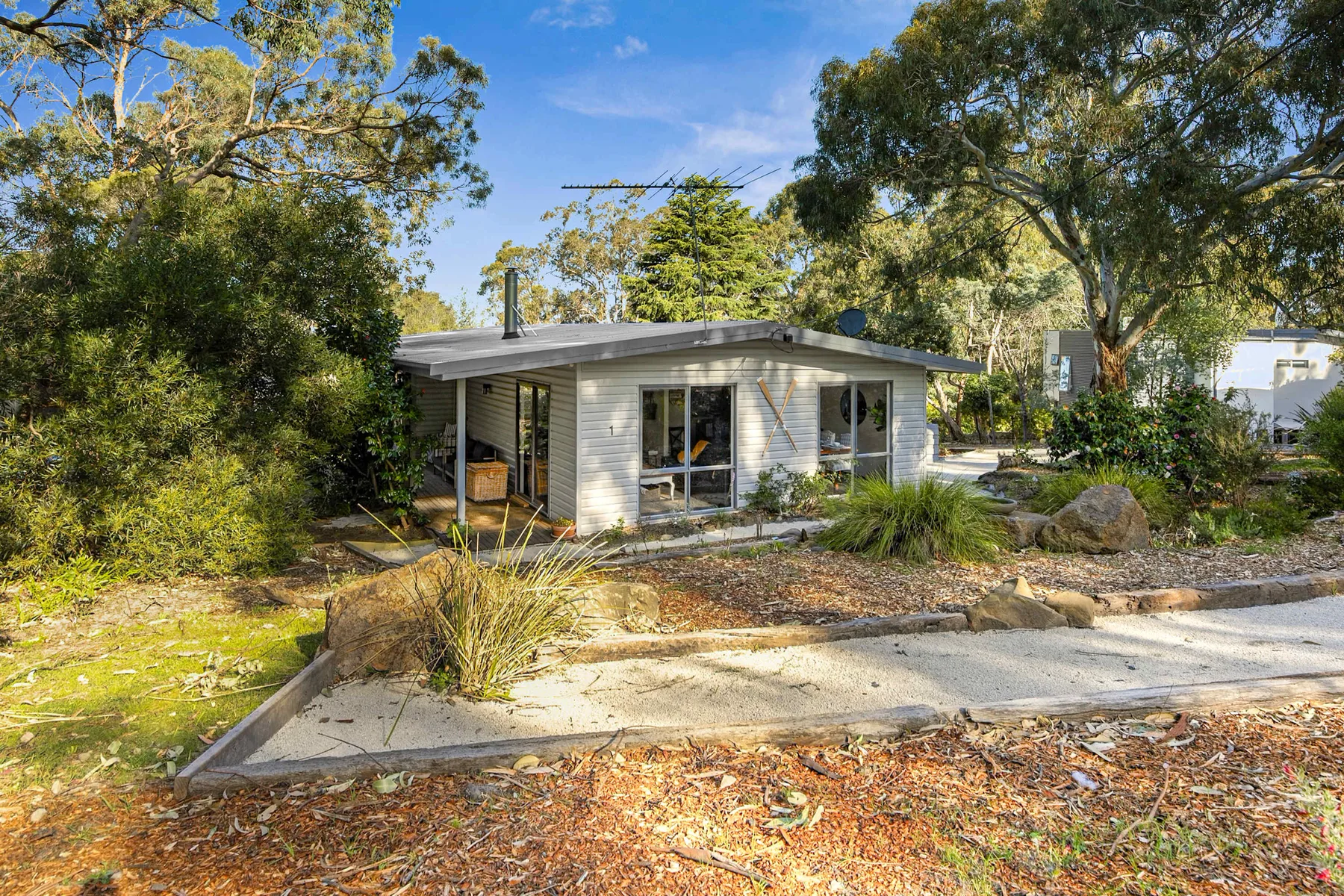 A small house sits among trees beside a paved path and landscaped garden.