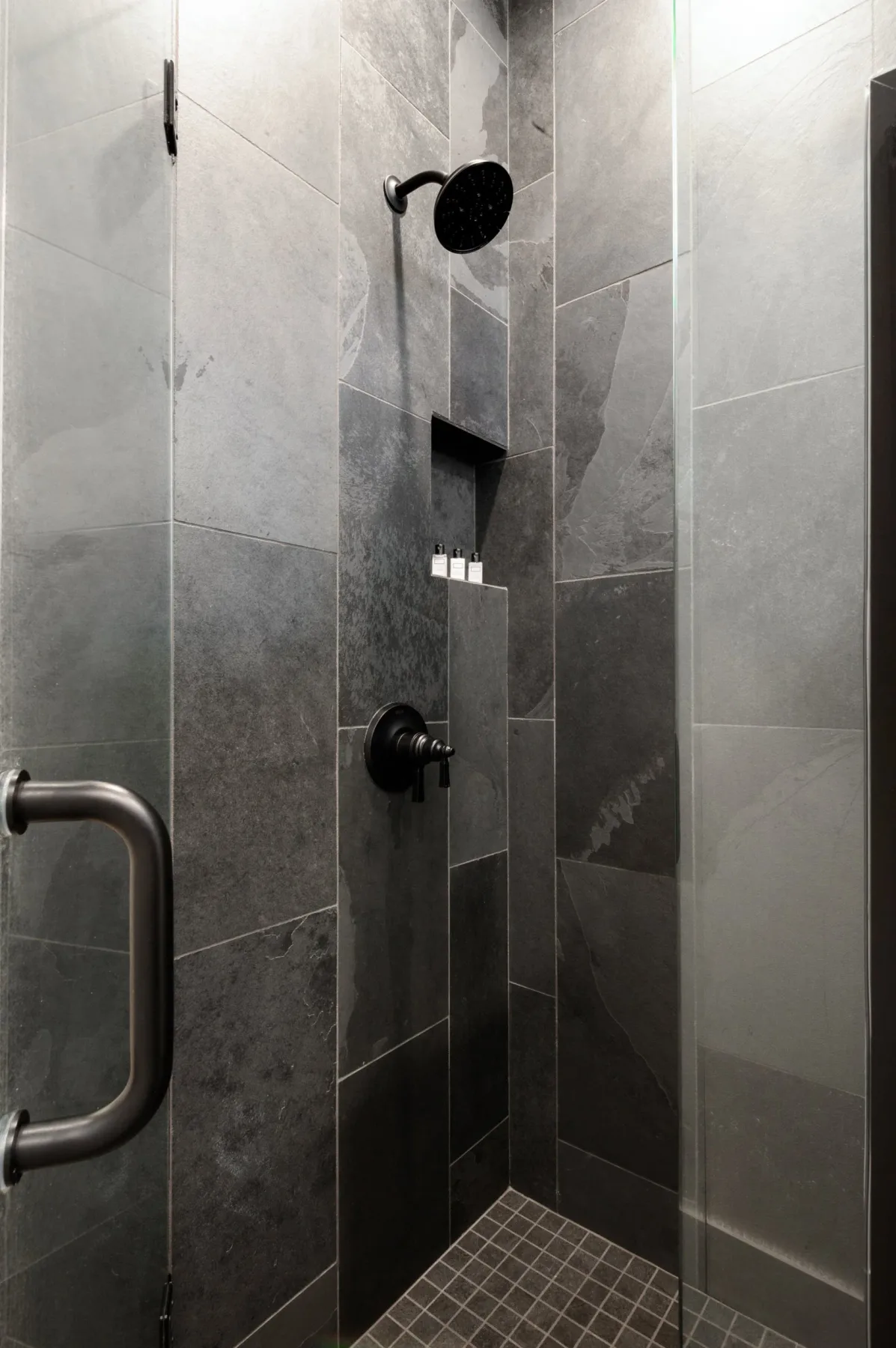 Representative hotel space: A tiled shower with a dark wall-mounted showerhead and recessed shelf.