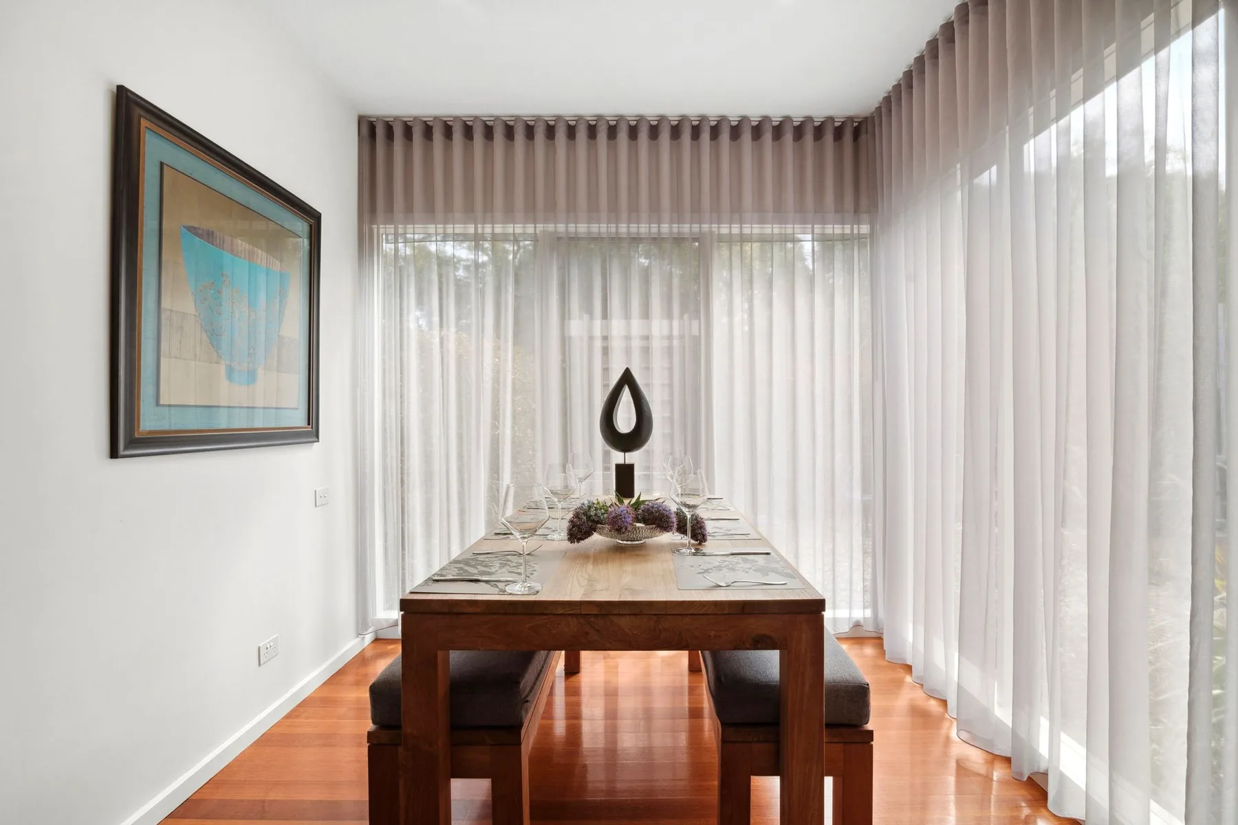 Una mesa de comedor está en una habitación luminosa con cortinas translúcidas y arte enmarcado.