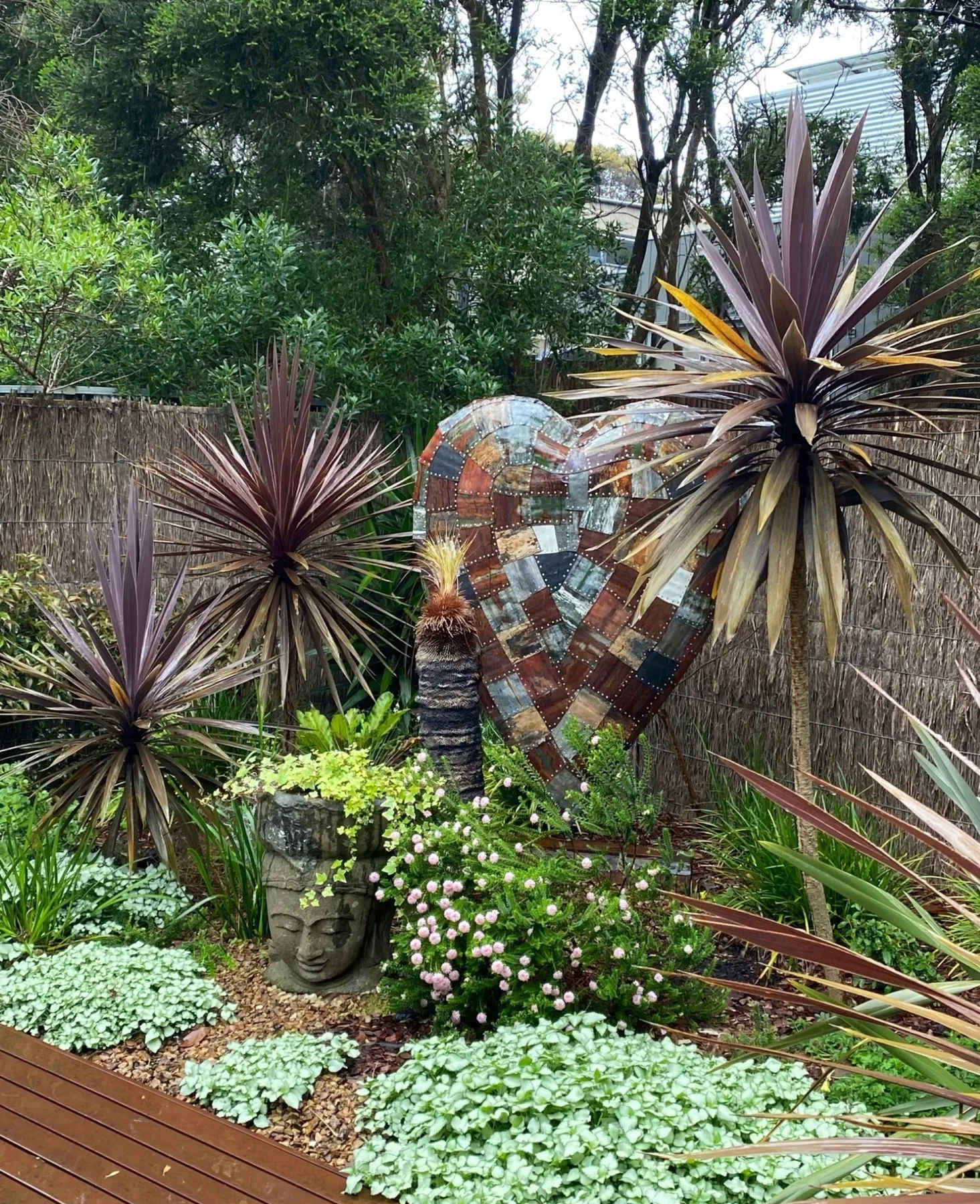 Un jardín con plantas puntiagudas, cubresuelo con flores y una escultura decorativa de mosaico.