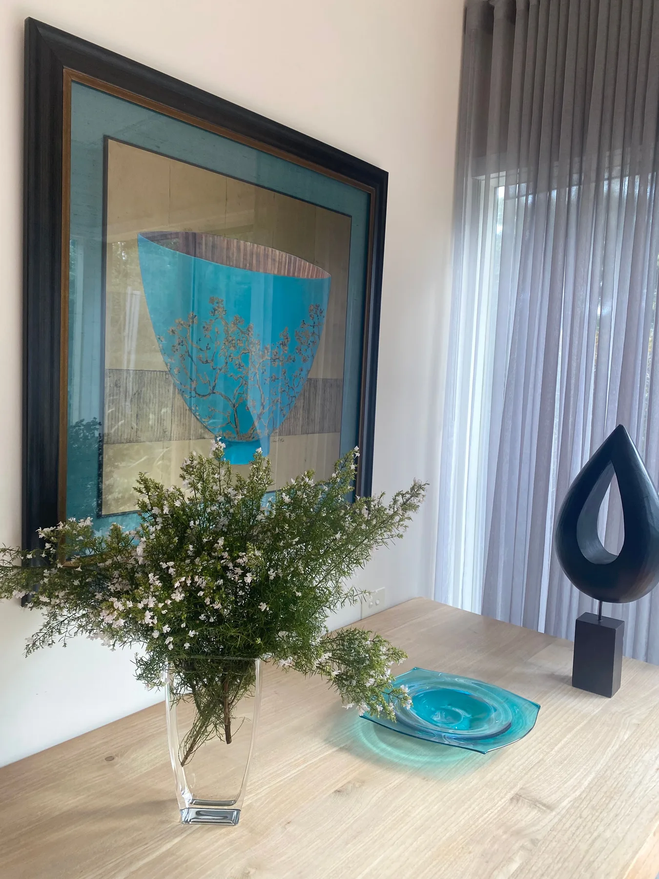 Una escena interior luminosa muestra una obra enmarcada, flores y un adorno escultórico.