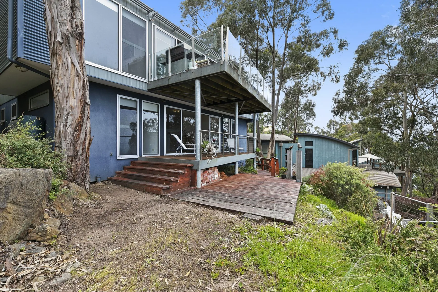 A modern house with a raised deck and large windows sits among trees.