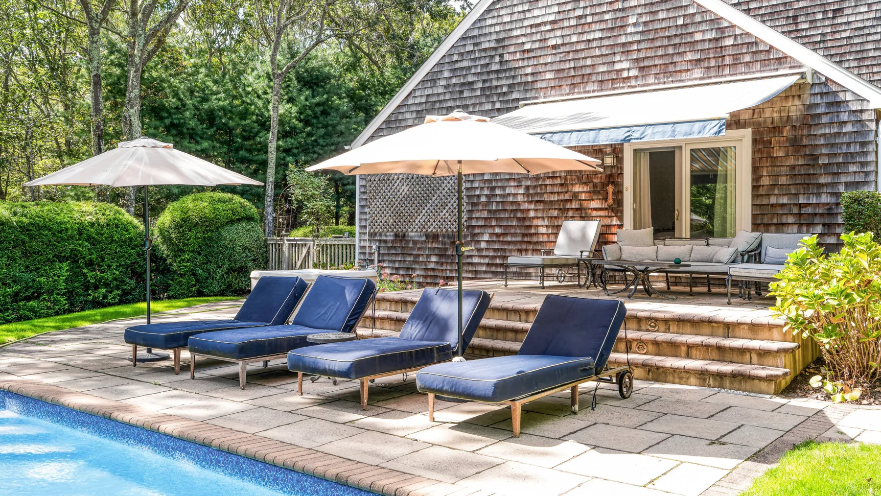 Un patio exterior junto a la piscina, con asientos reclinables y sombrillas junto a una casa con revestimiento de tejas.