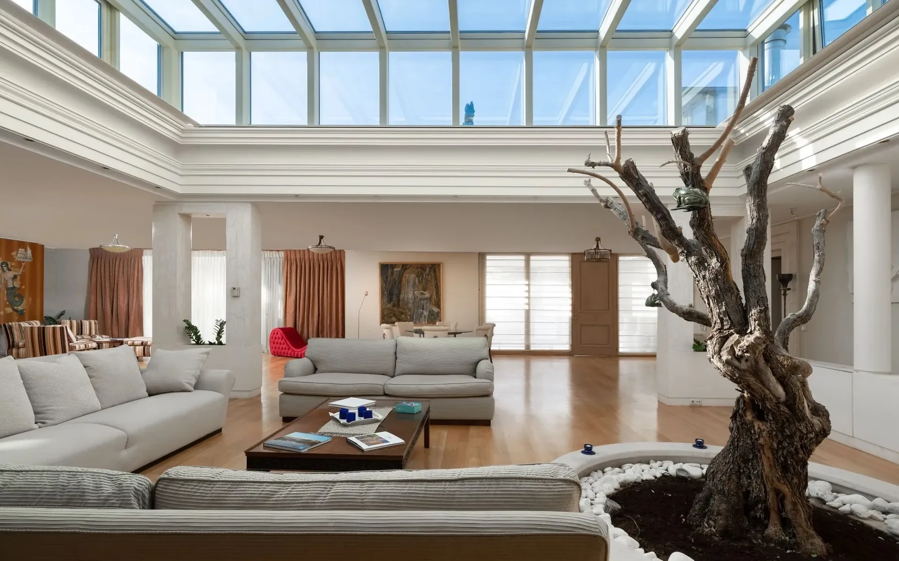 A bright living room with seating beneath a glass ceiling and a sculptural indoor tree.