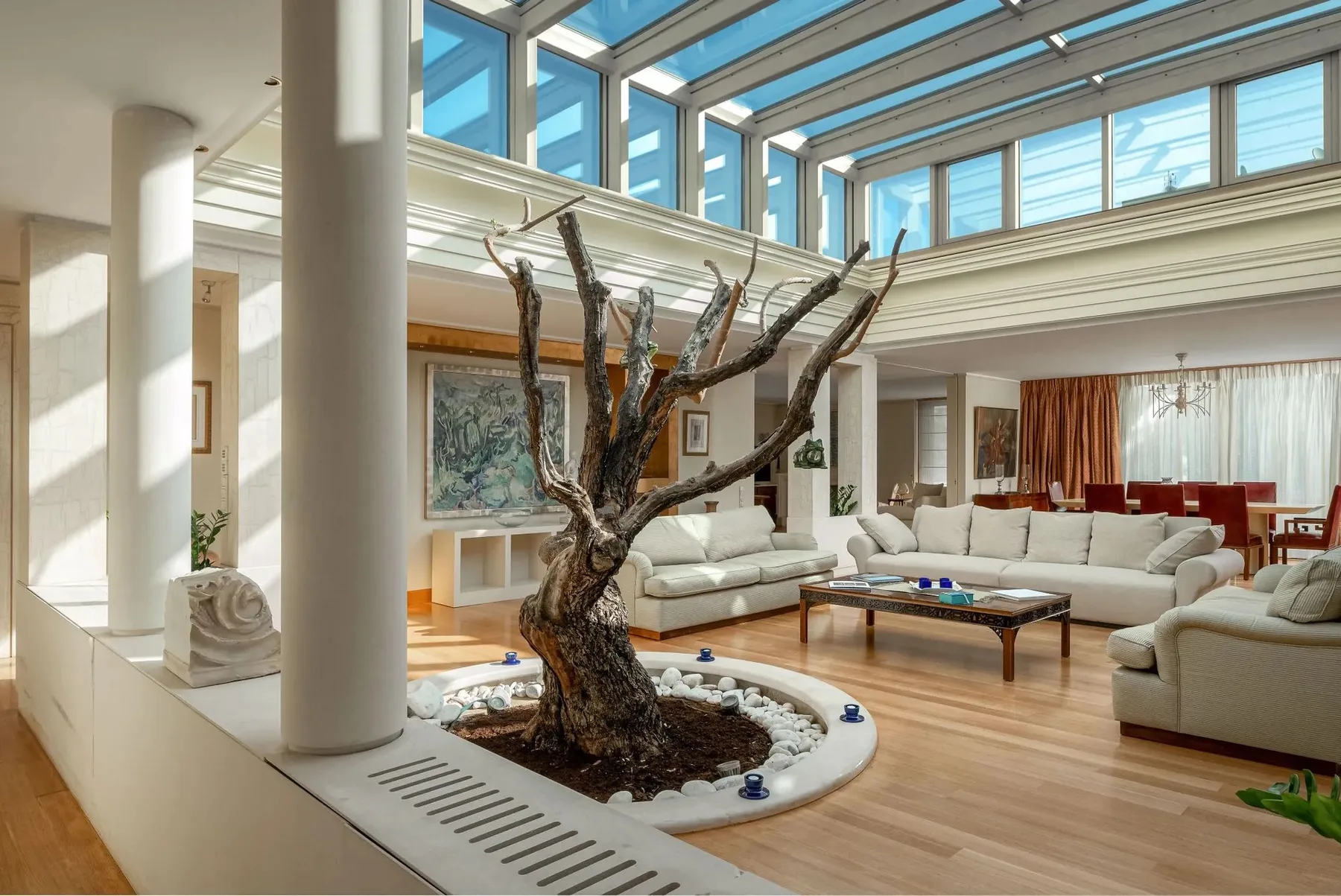 A bright living room has large windows, seating, and a sculptural tree centerpiece.