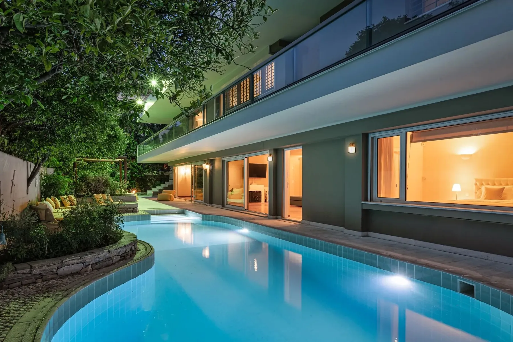 A modern house overlooks a lit swimming pool and landscaped garden at night.