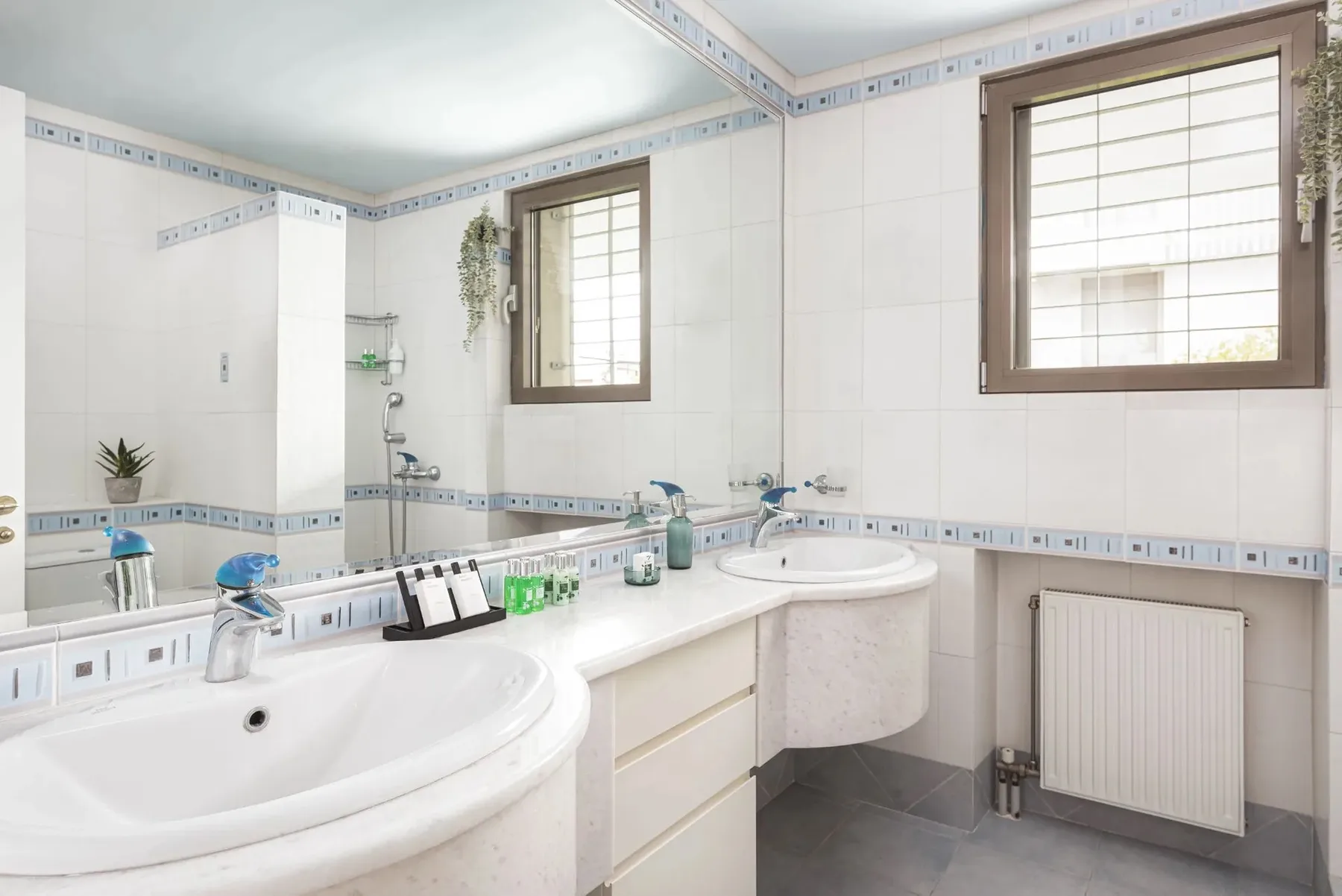 A tiled bathroom with two sinks, mirrors, and a radiator beneath a window.