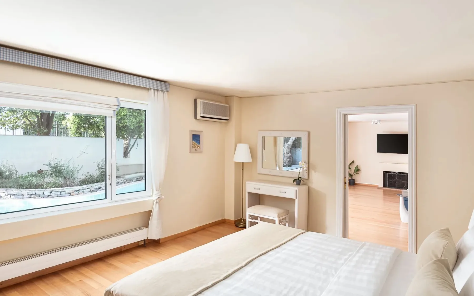 A bright bedroom with a bed, vanity, and large windows overlooking greenery.