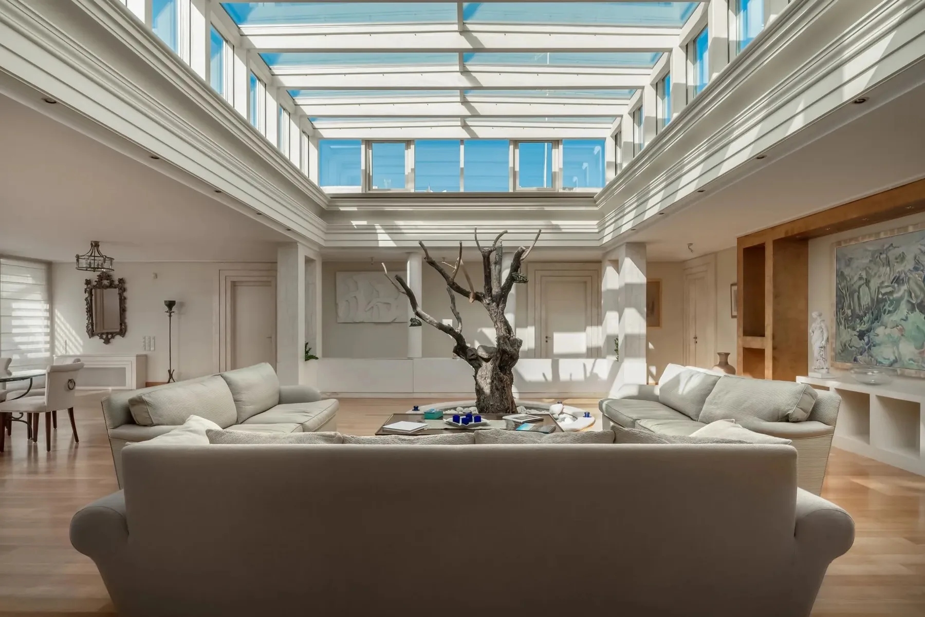 A bright living room with seating arranged around a central table beneath a glass ceiling.