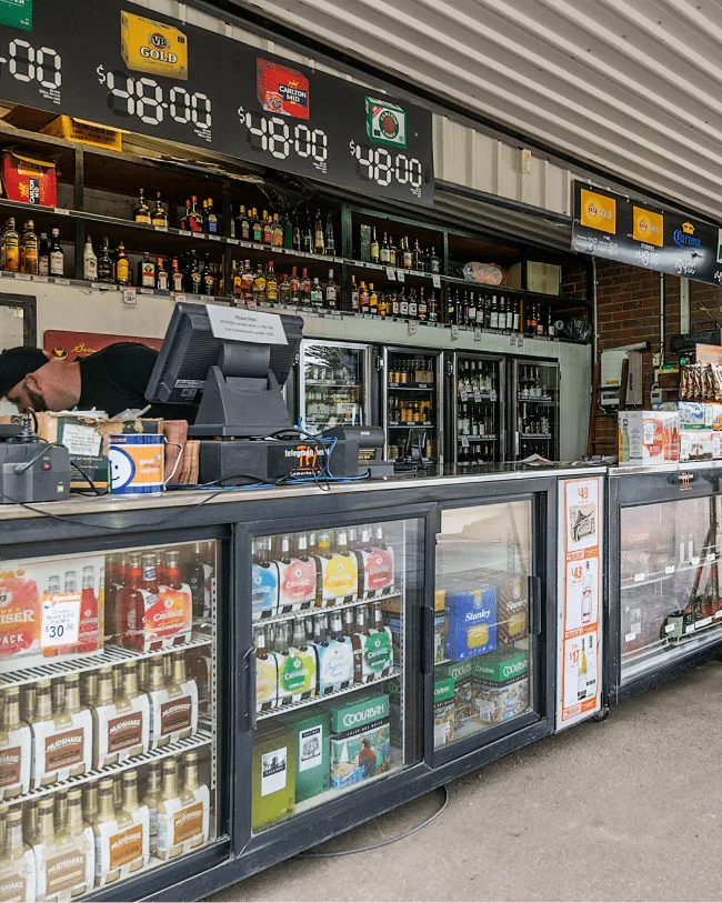 Espacio representativo del hotel: Un mostrador de tienda exhibe bebidas en vitrinas refrigeradas bajo estantes con botellas.