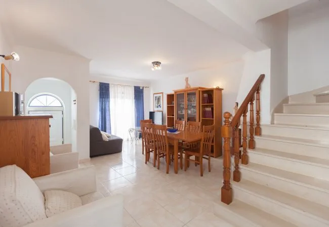 A bright dining and living area with a wooden dining table and staircase.