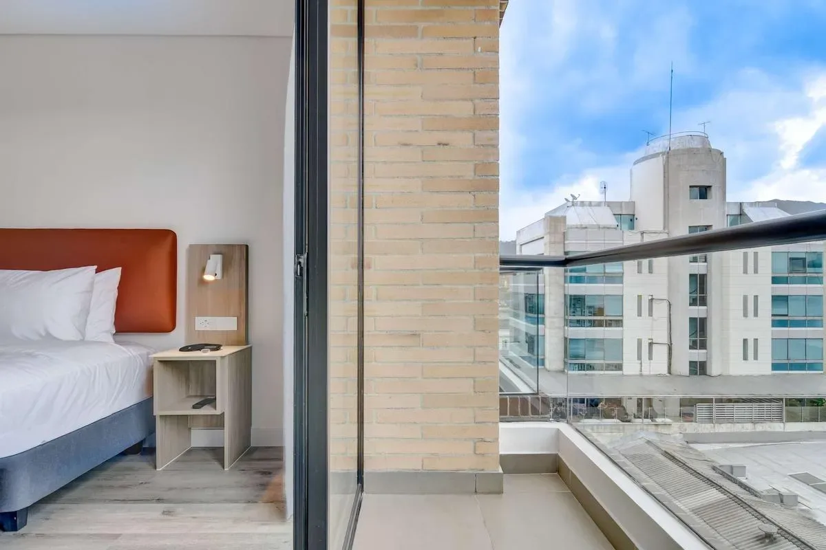 Un dormitorio da a un balcón, con una cama bien hecha y edificios modernos visibles al exterior.