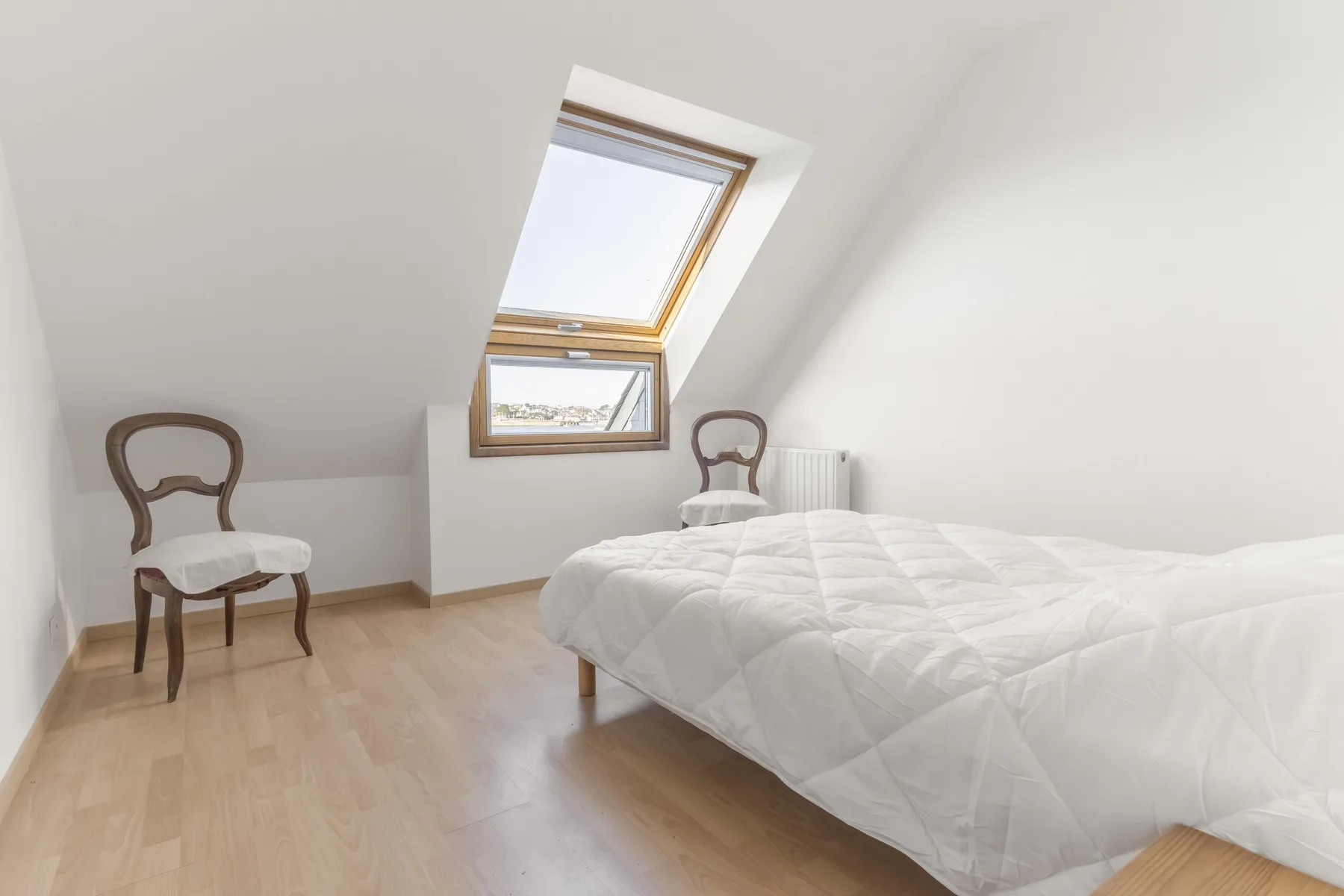 A bright attic bedroom with a bed, two chairs, and slanted ceilings.