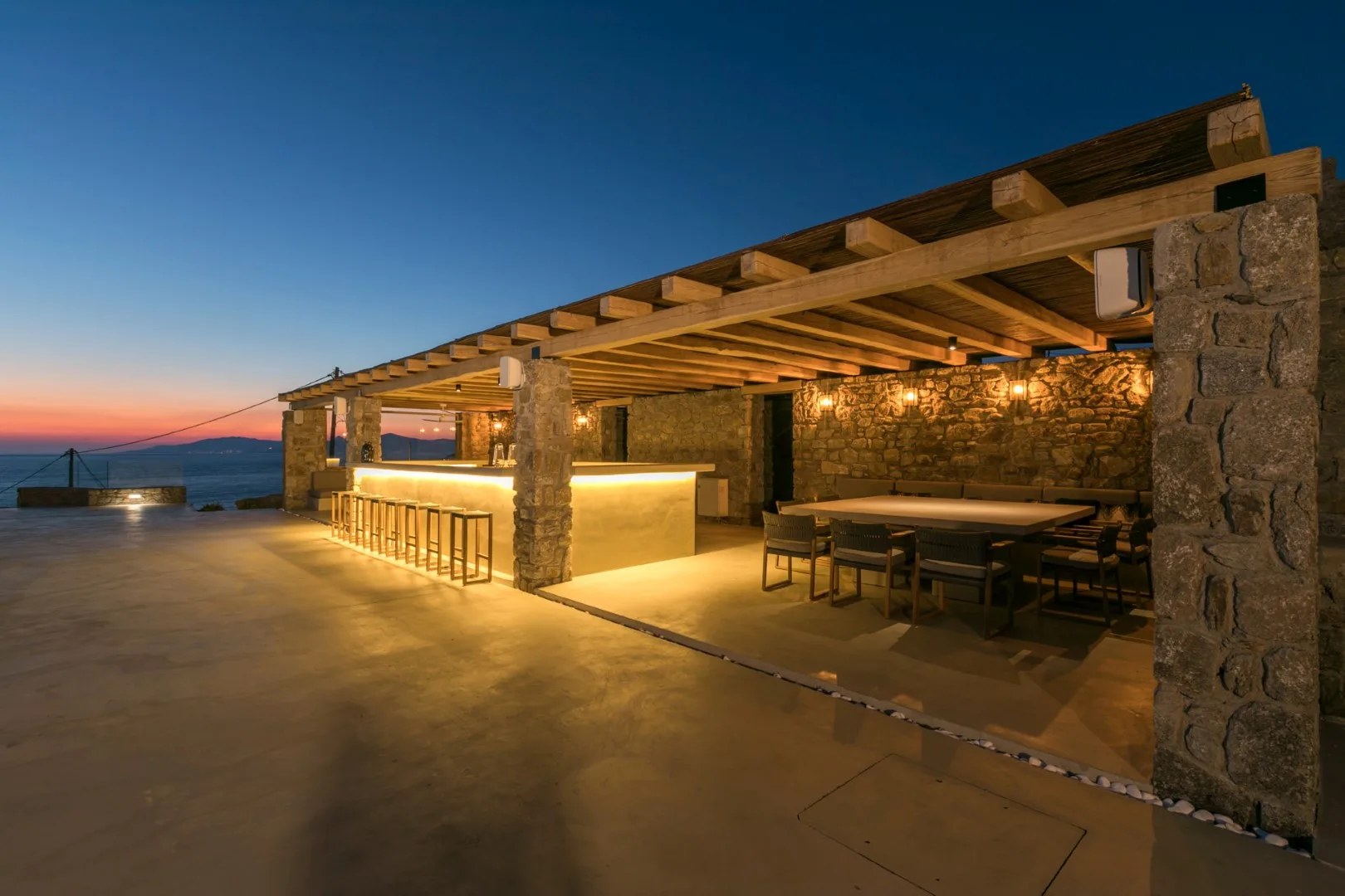 A covered outdoor dining area with stone walls and a view of the sea at sunset.