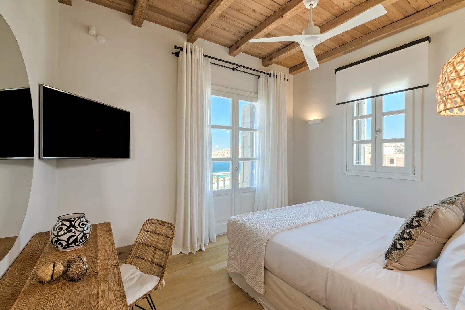A bright bedroom with a bed, a wall-mounted television, and windows.
