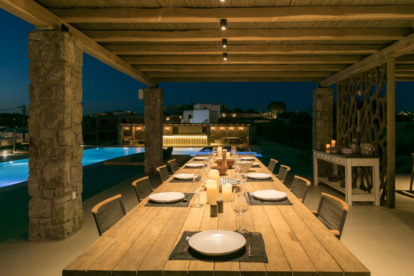An outdoor dining table is set beneath a wooden pergola beside a swimming pool.