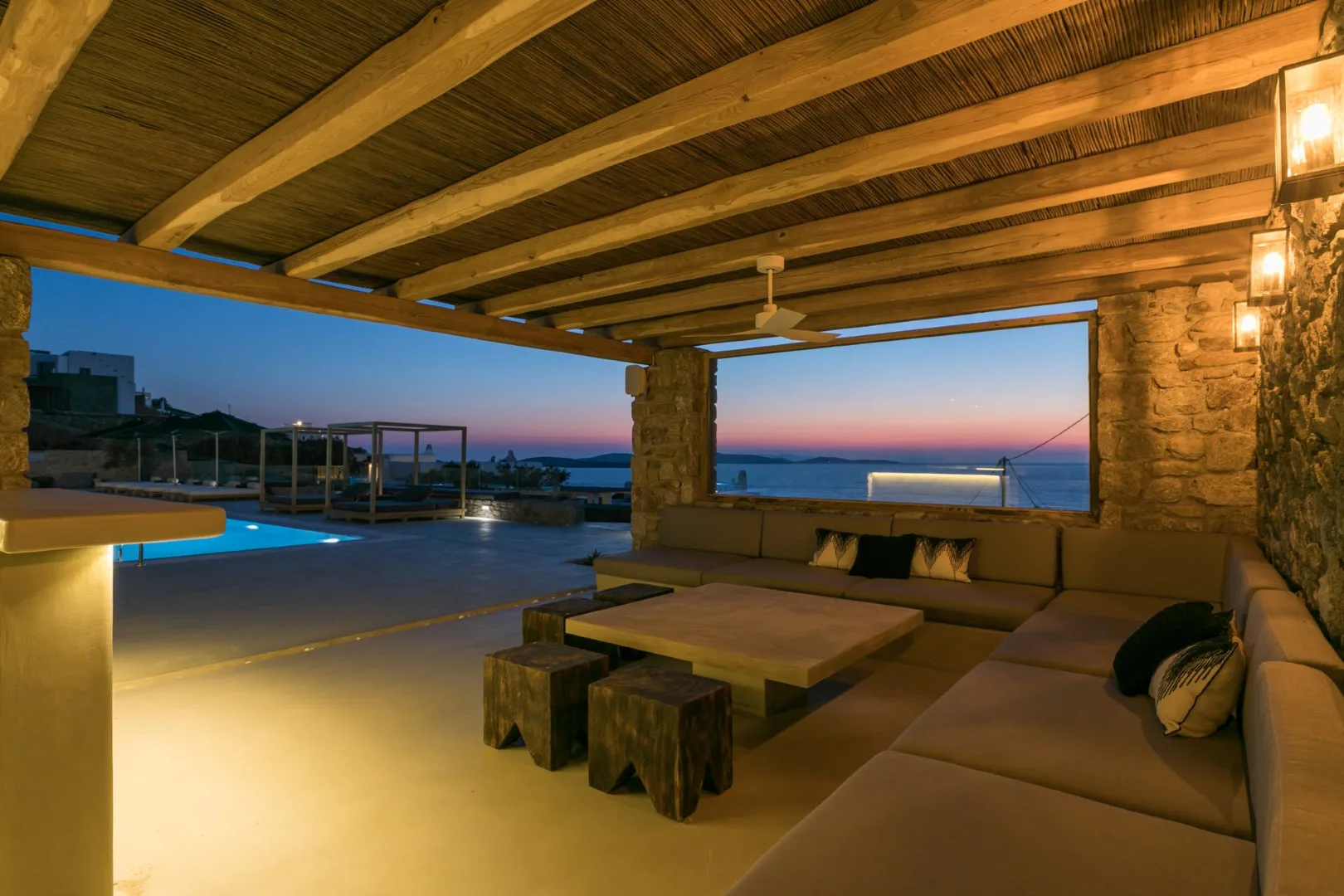 A covered outdoor lounge with cushioned seating overlooks a pool and the sea.