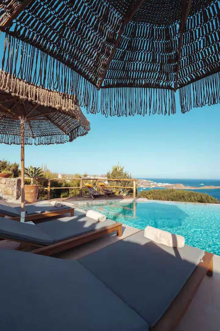 Una zona de descanso junto a la piscina al aire libre mira al mar bajo sombrillas con flecos.