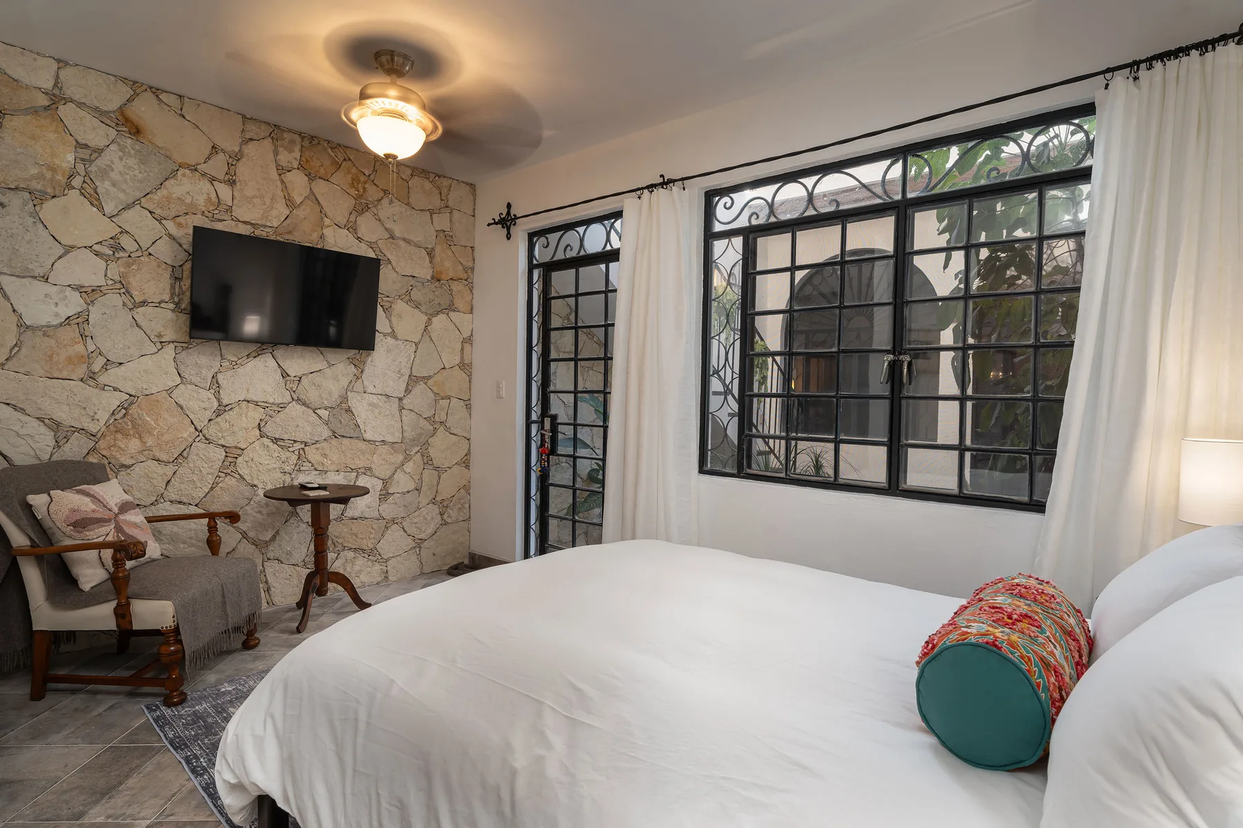 A bedroom with a white bed, stone accent wall, and large black-framed windows.