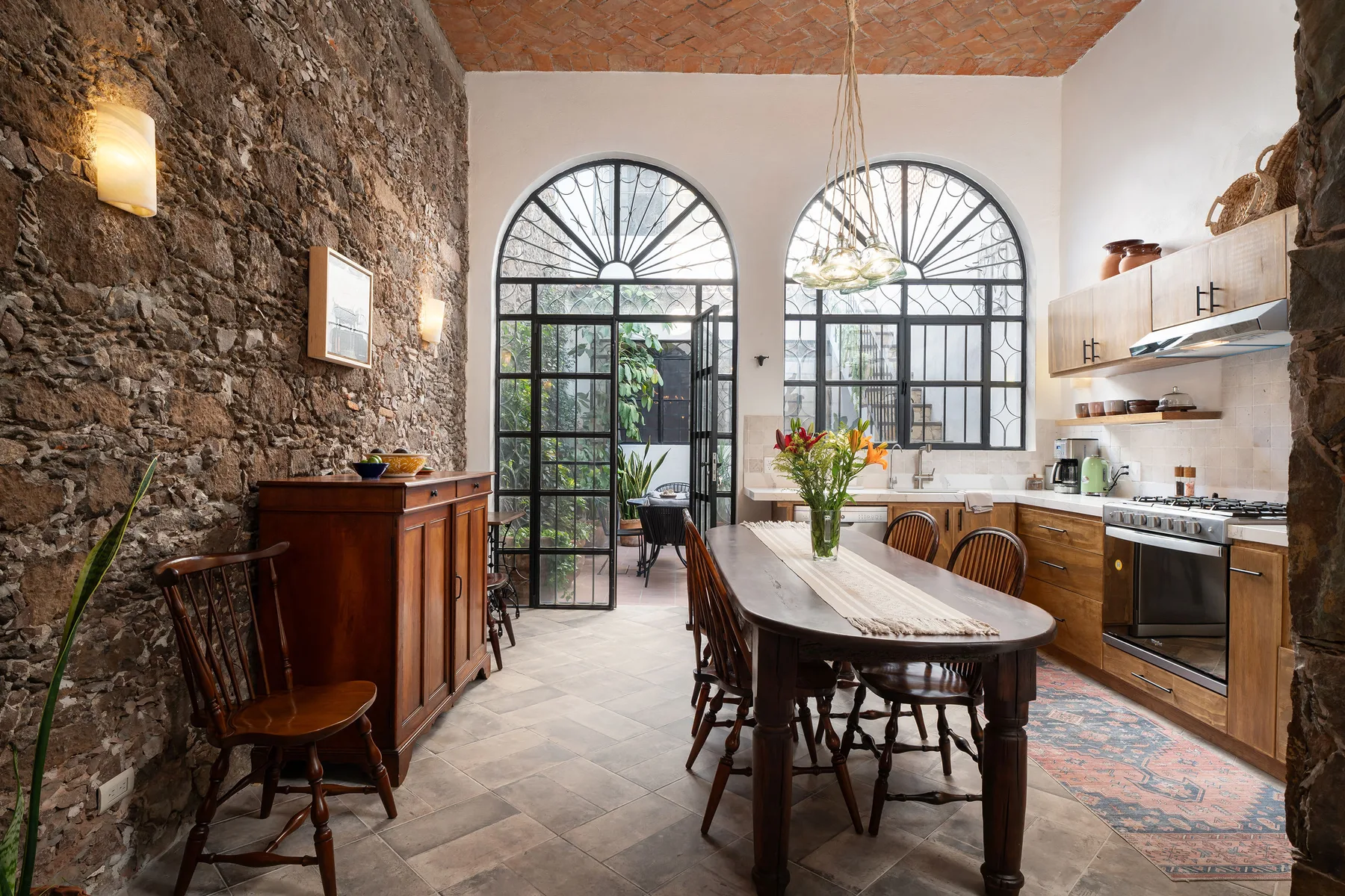 A rustic kitchen and dining area with a long table beneath arched windows.