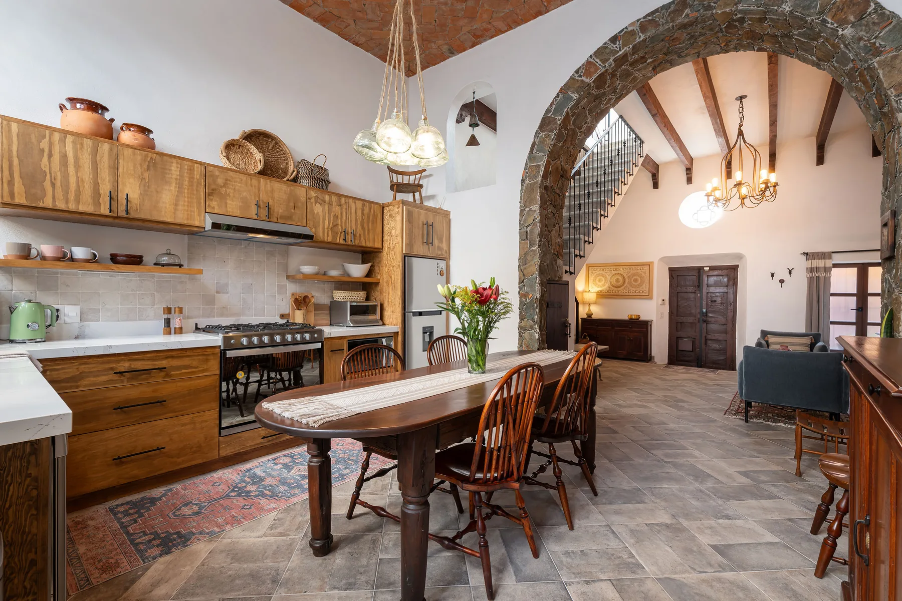 A rustic kitchen and dining area with a long wooden table beneath a stone arch.