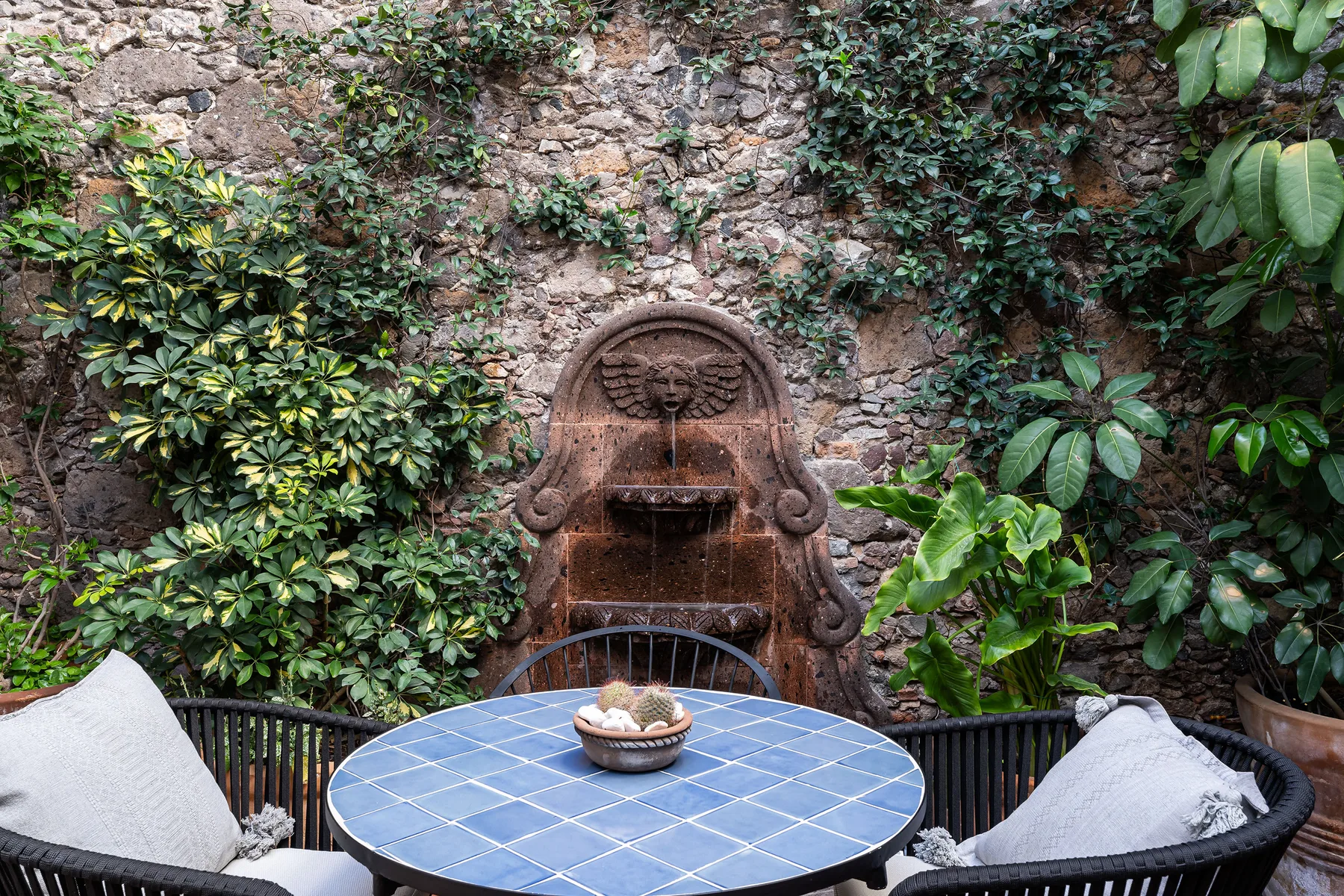 A garden patio with a blue-tiled table and a wall fountain surrounded by greenery.