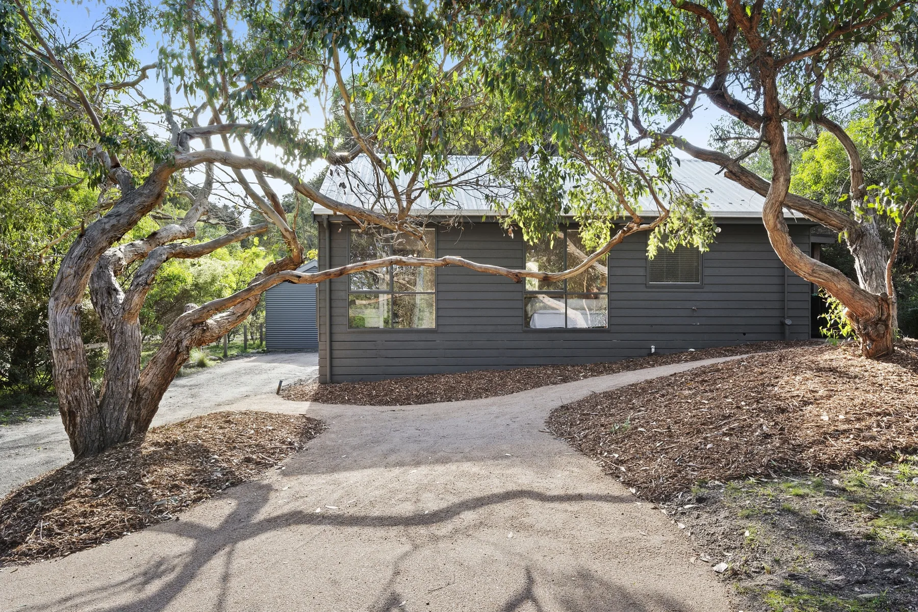 A dark-sided house stands among leafy trees beside a paved path.