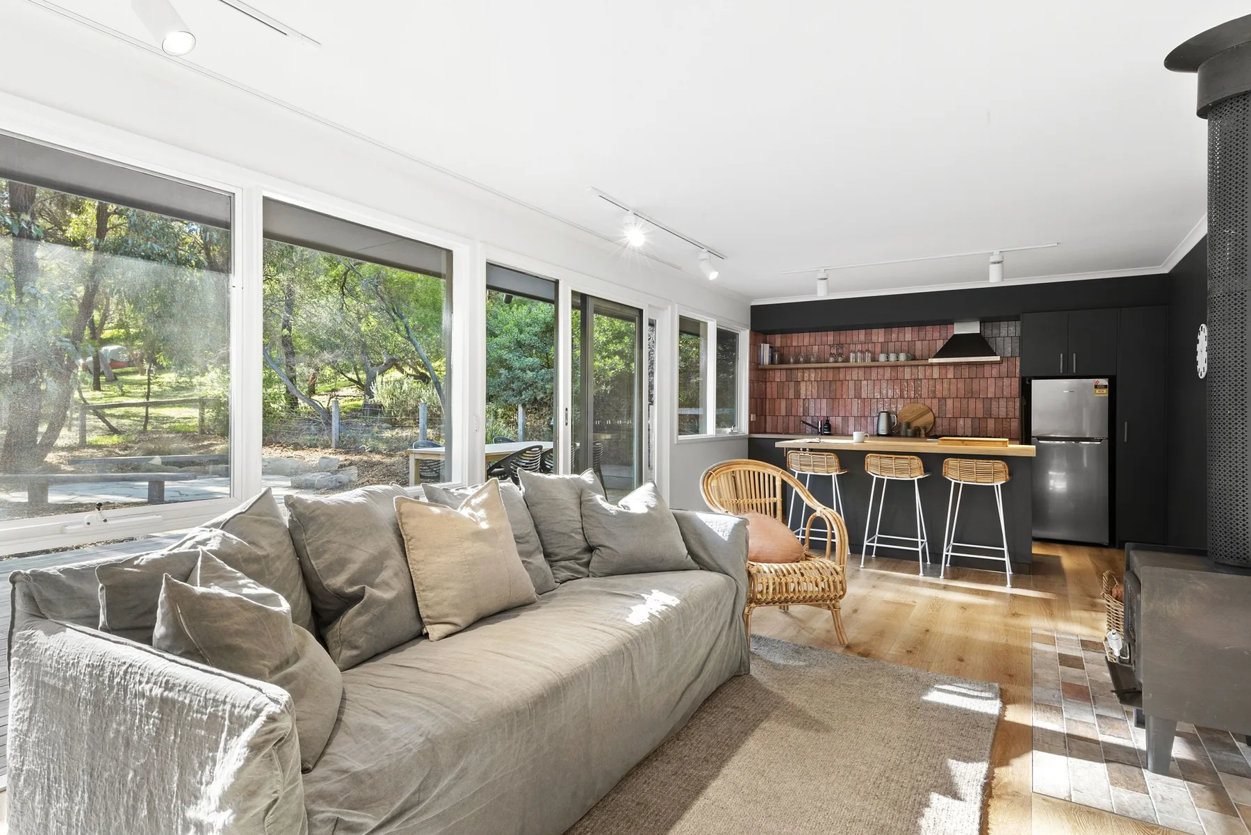 A bright living room opens onto a kitchen, with large windows overlooking trees.