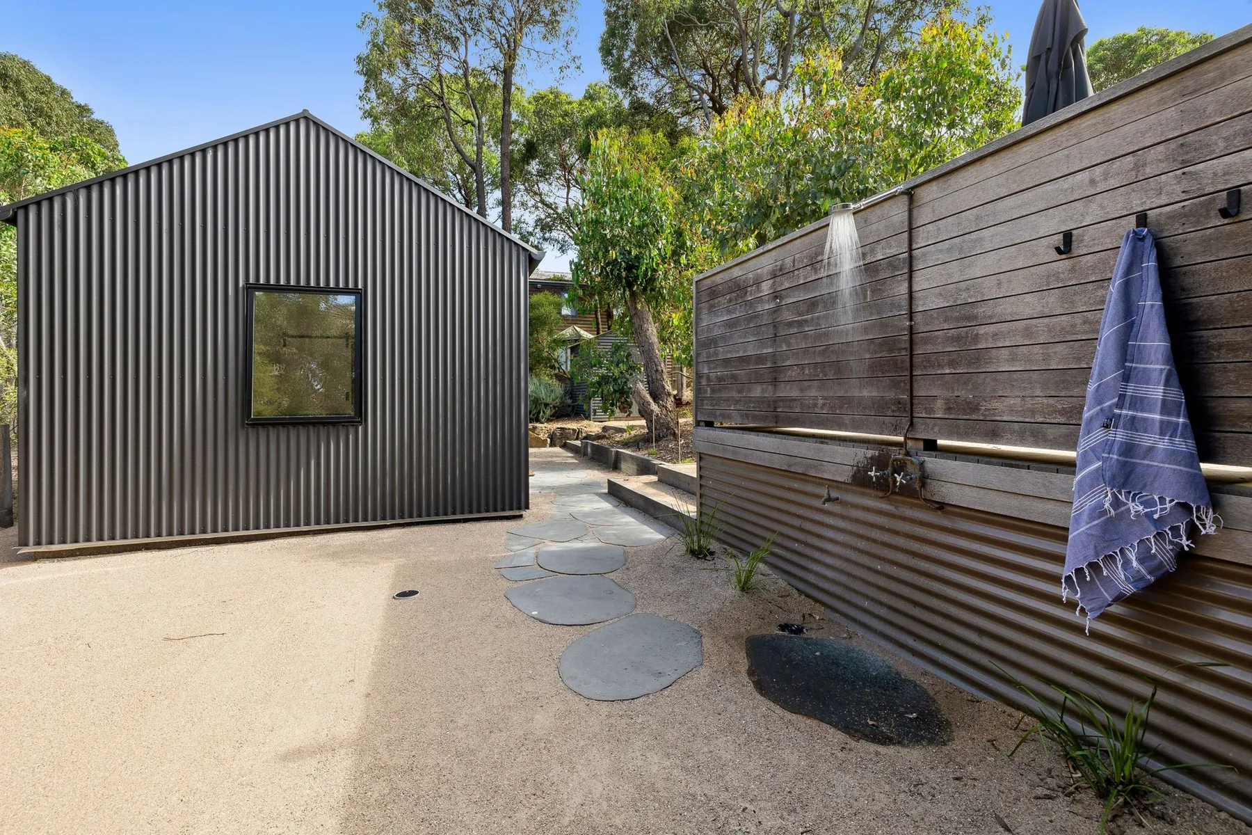 An outdoor shower area beside a dark shed, with stepping stones across the sandy ground.