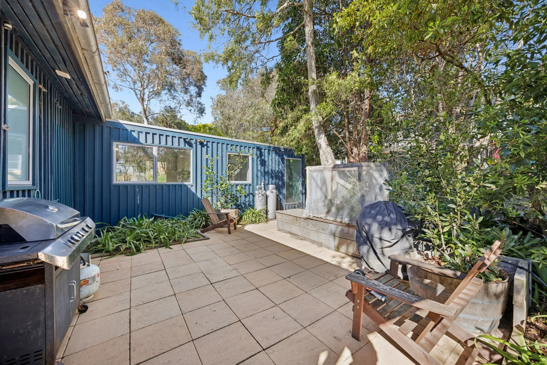 A paved outdoor patio beside a blue building, with seating and surrounding trees.