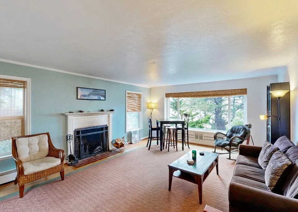 A bright living room has seating, a fireplace, and a dining table beside large windows.