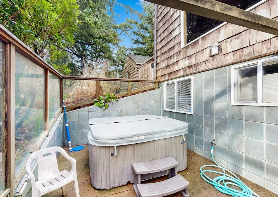 An outdoor hot tub sits beside a tiled wall, with a garden hose nearby.