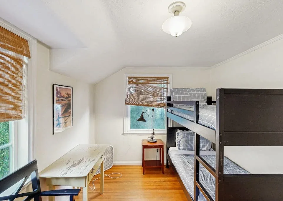 A bedroom with bunk beds, a desk, and windows with woven blinds.