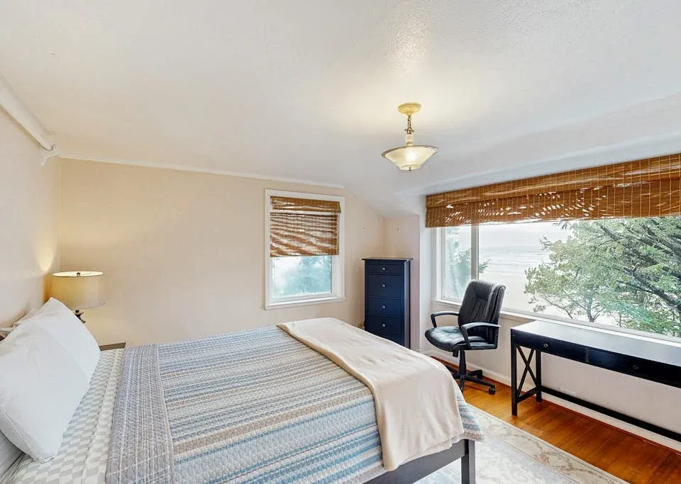 A bedroom with a bed and a desk beside a large window.