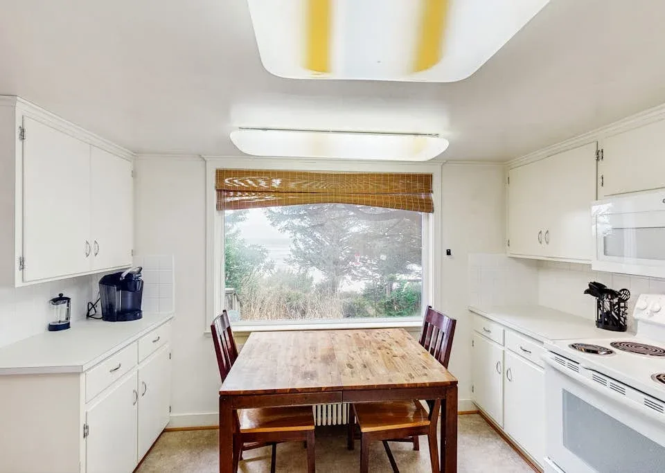 A bright kitchen with white cabinets, a wooden dining table, and a large window.