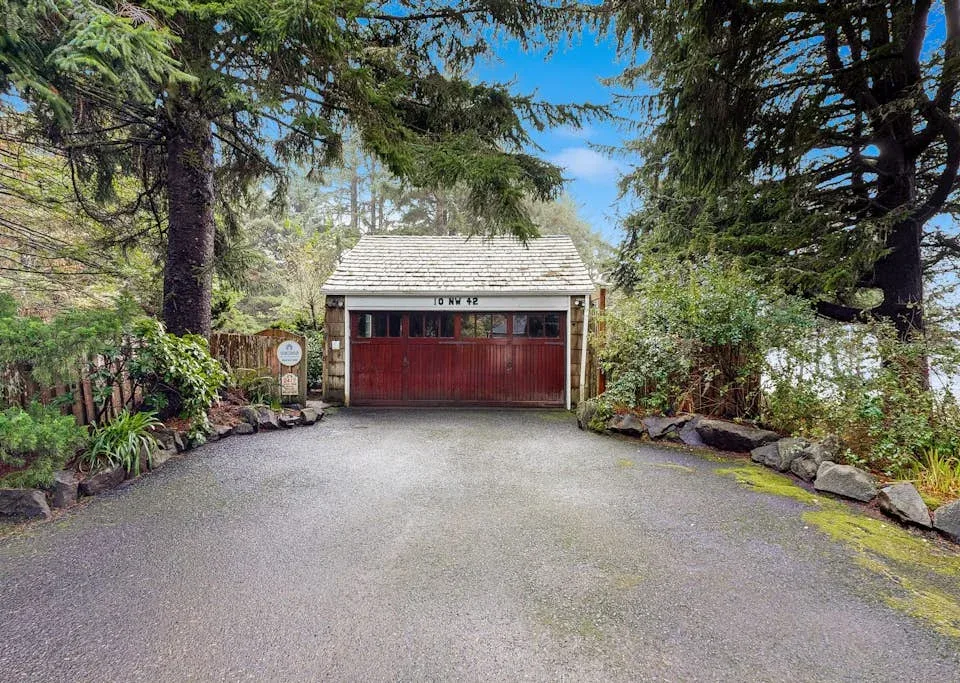 A red garage sits at the end of a paved driveway beneath tall trees.