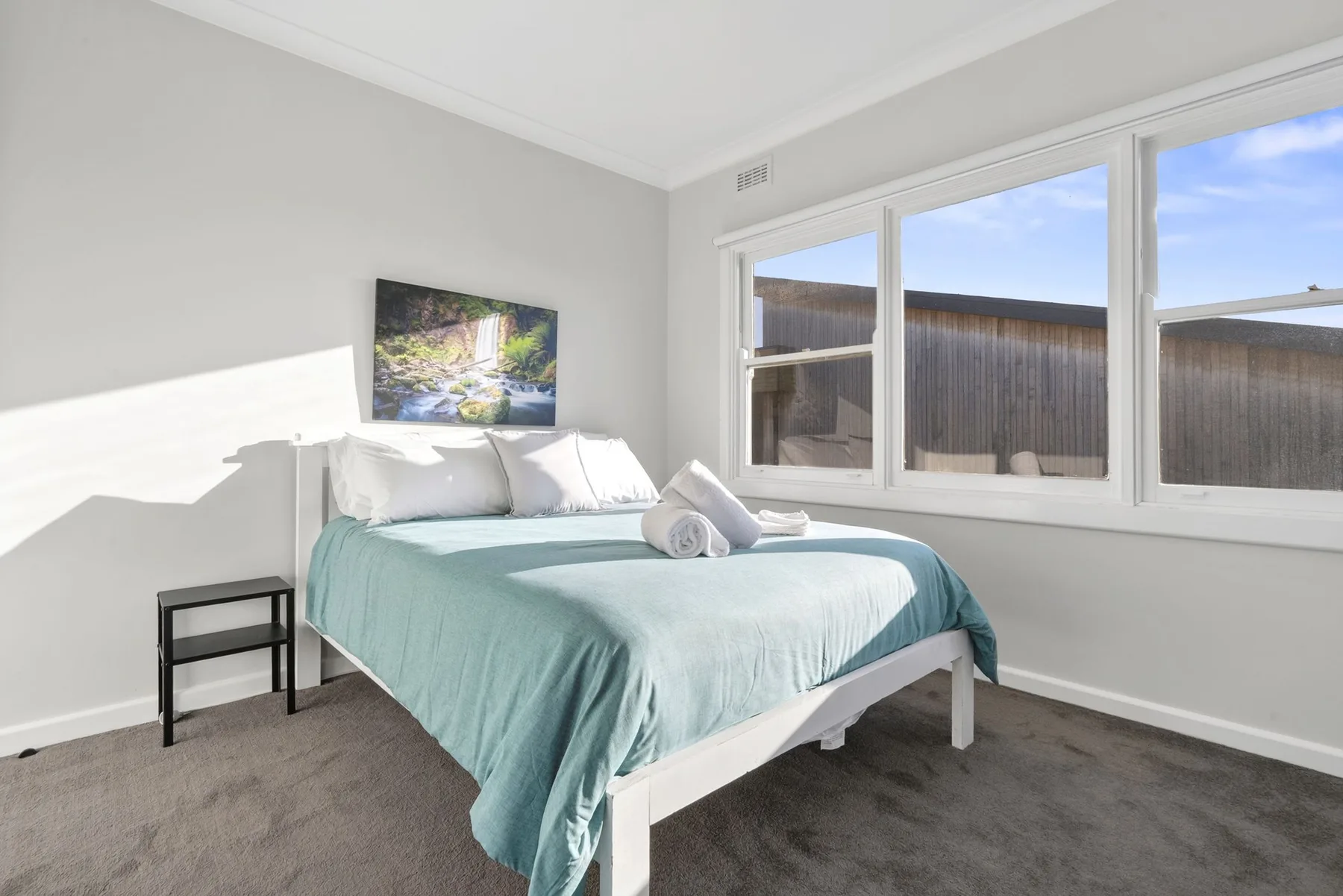 A bright bedroom with a bed dressed in pale teal bedding beside wide windows.