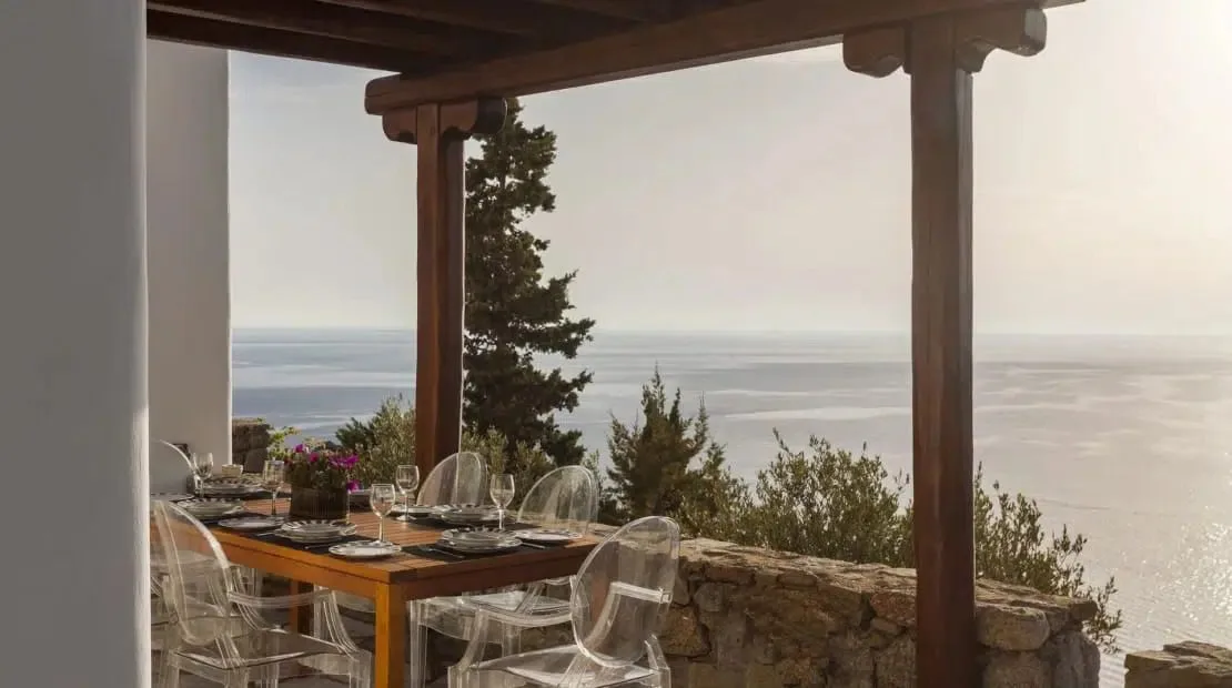 An outdoor dining area under a wooden pergola overlooks the sea.
