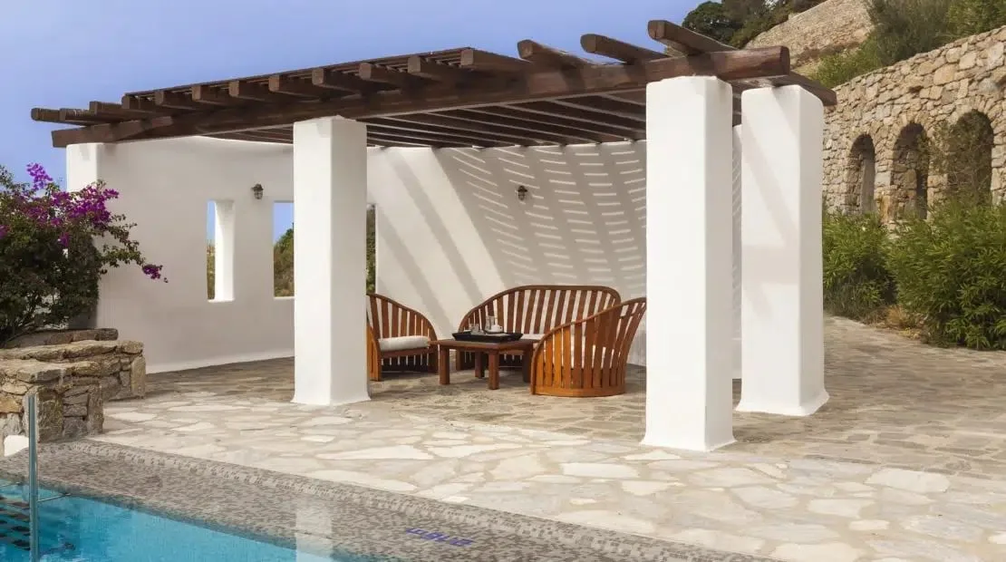 A shaded patio with curved wooden seating beside a swimming pool.