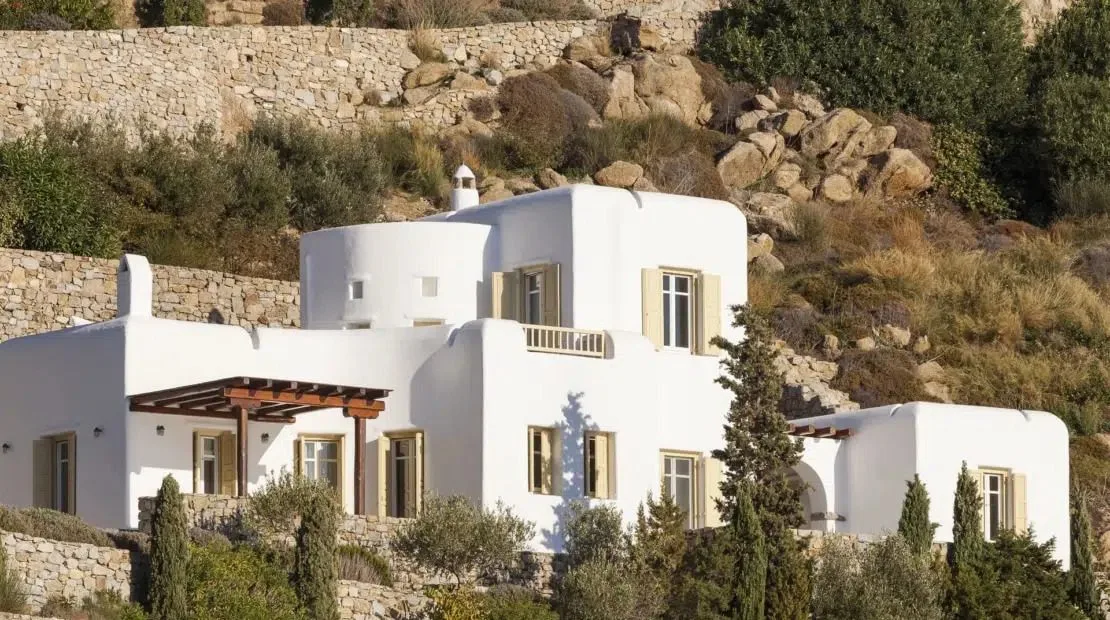A white building with rounded walls sits among shrubs below a rocky hillside.