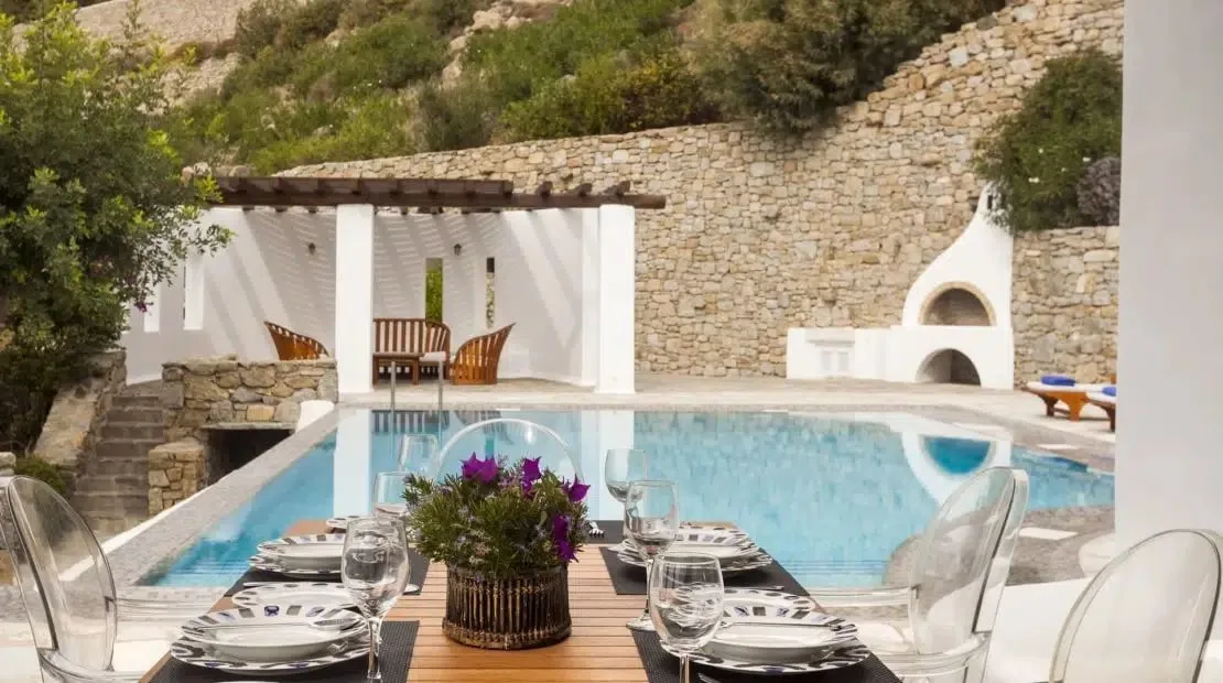 A dining table beside a swimming pool, with stone walls and an outdoor fireplace.