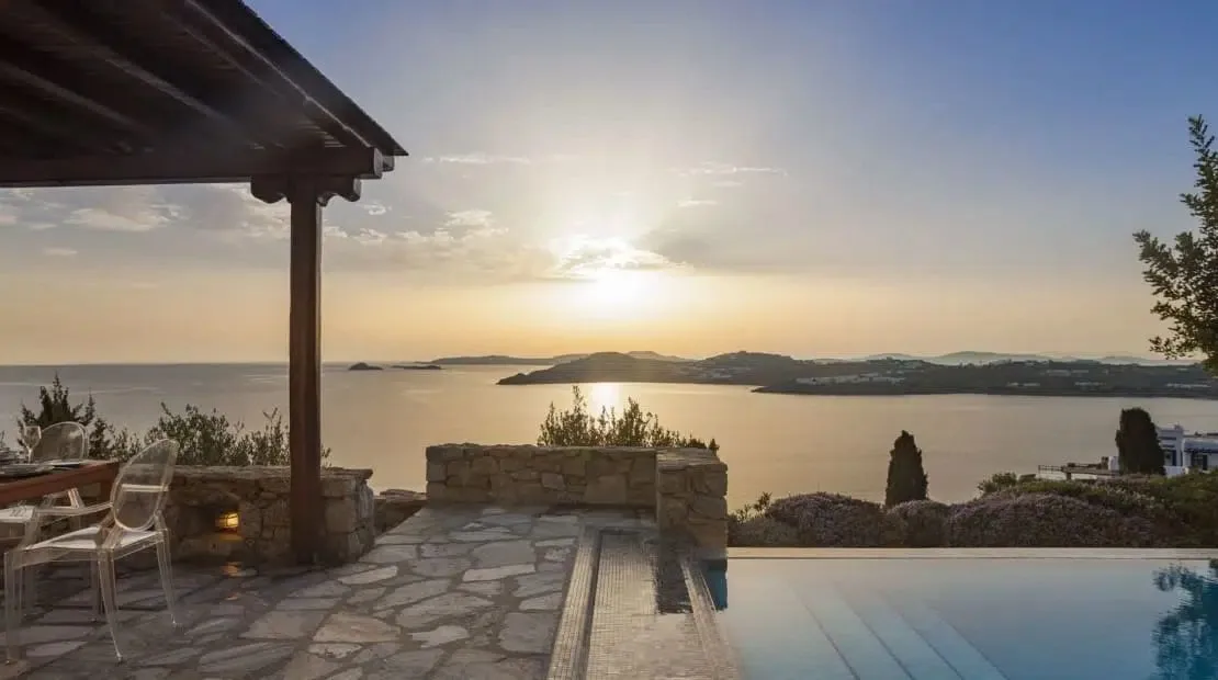 A stone patio overlooks calm water and distant islands at sunset.