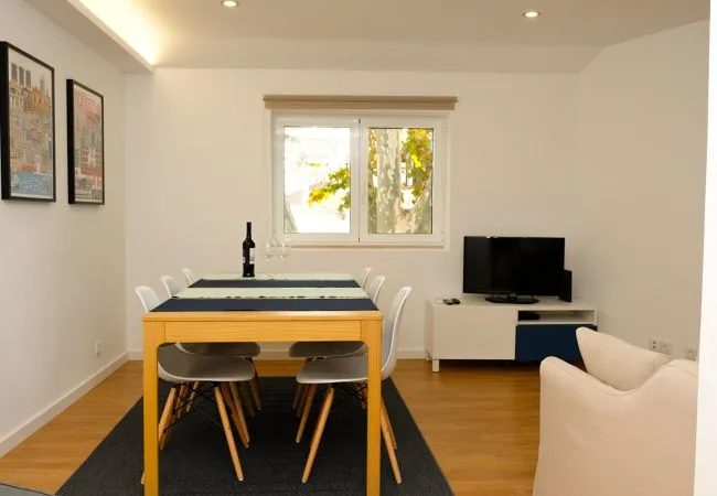 A dining and living area with a table, seating, and a television by the window.