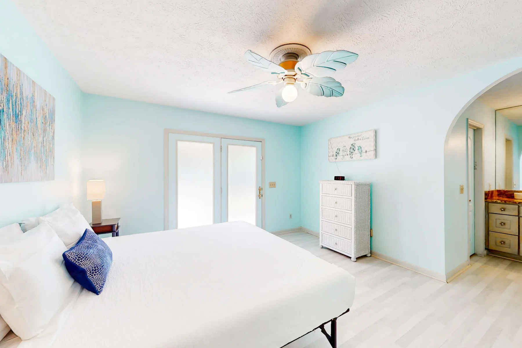 A bright bedroom with a large bed, pale blue walls, and a ceiling fan.
