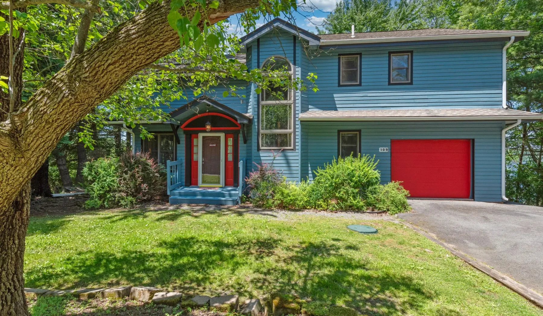 Una casa azul de dos plantas con una puerta de garaje roja está rodeada de árboles frondosos.