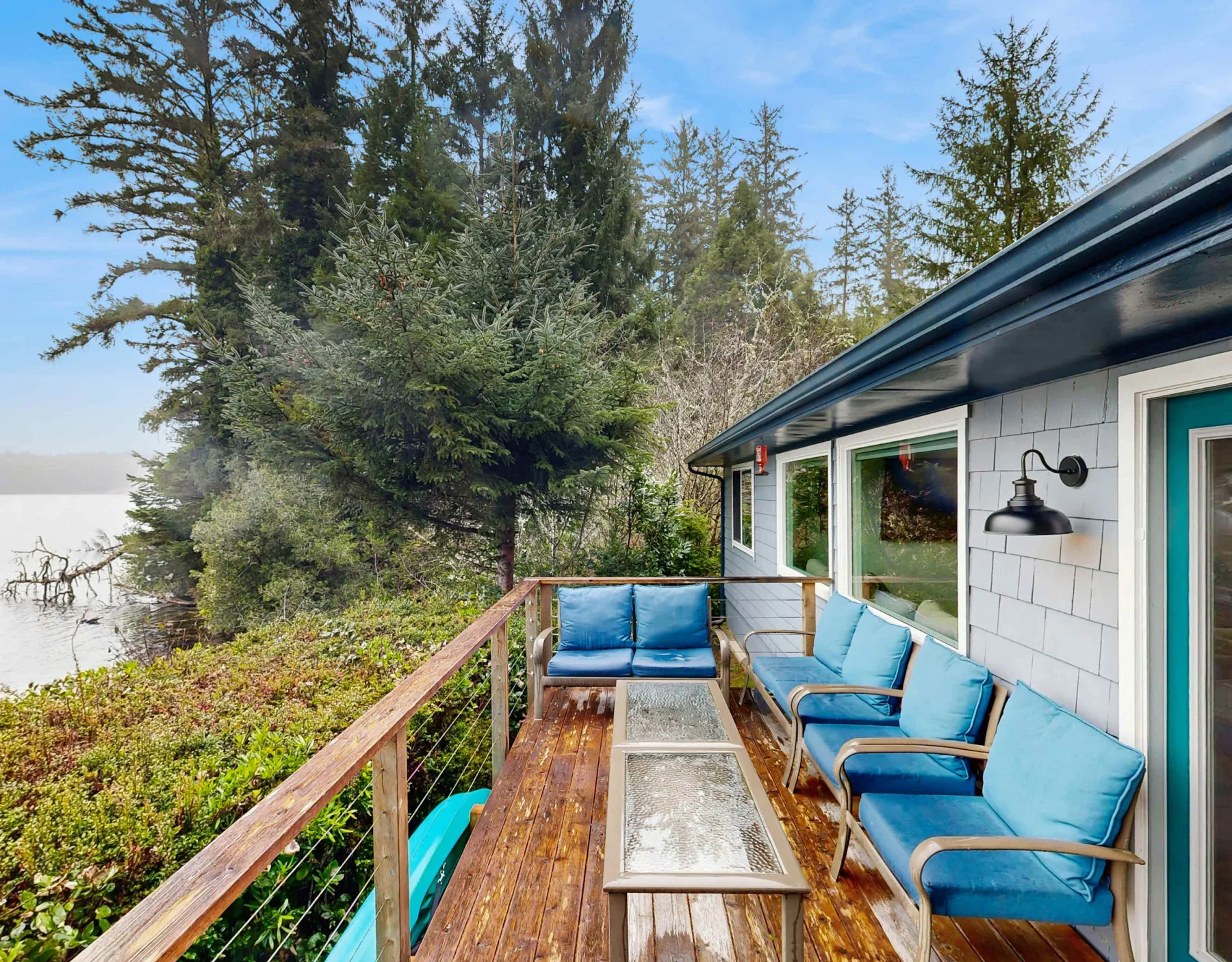 Una terraza de madera junto a la casa tiene asientos azules y una mesa con superficie de vidrio.