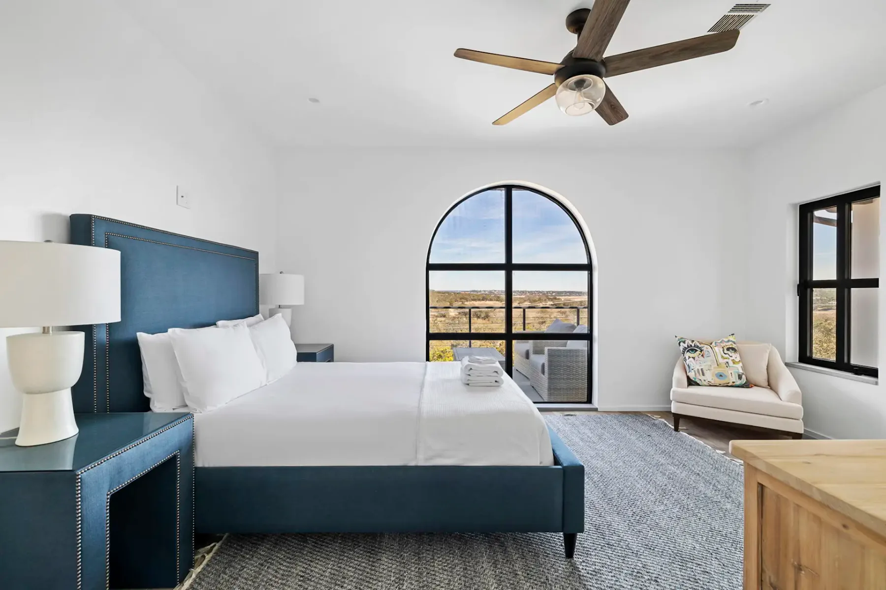 A bright bedroom with a blue bed and an arched window overlooking open land.