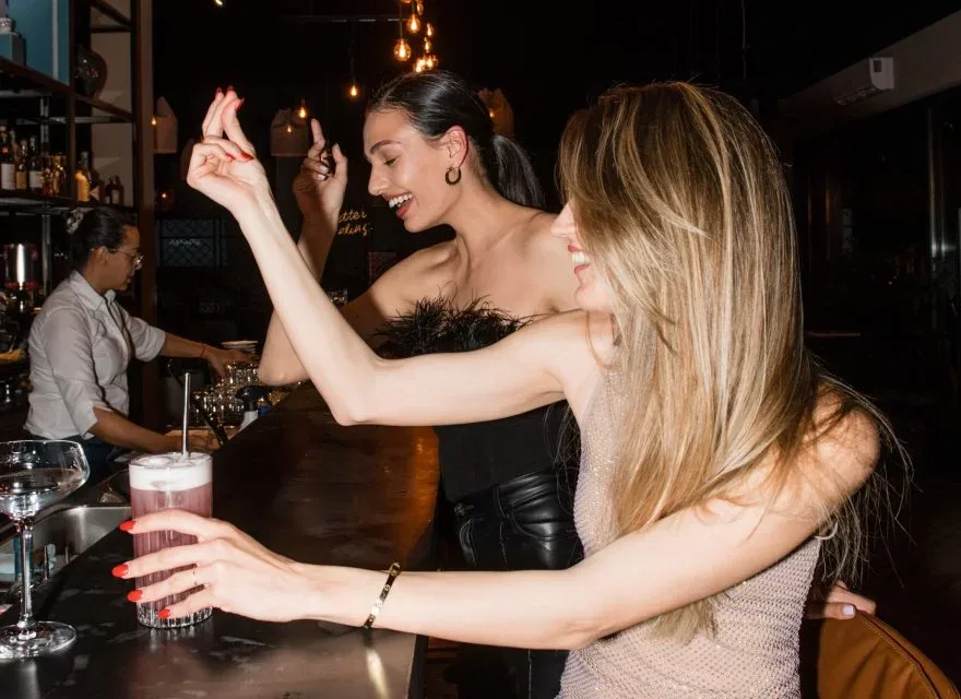 Espacio representativo del hotel: Mujeres bailan junto a una barra mientras un camarero sirve bebidas en un bar con poca luz.