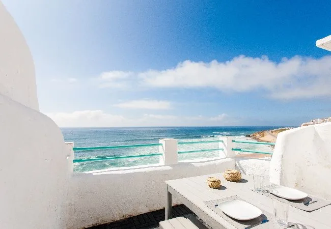 A seaside terrace overlooks the ocean, with a table and seating in the foreground.